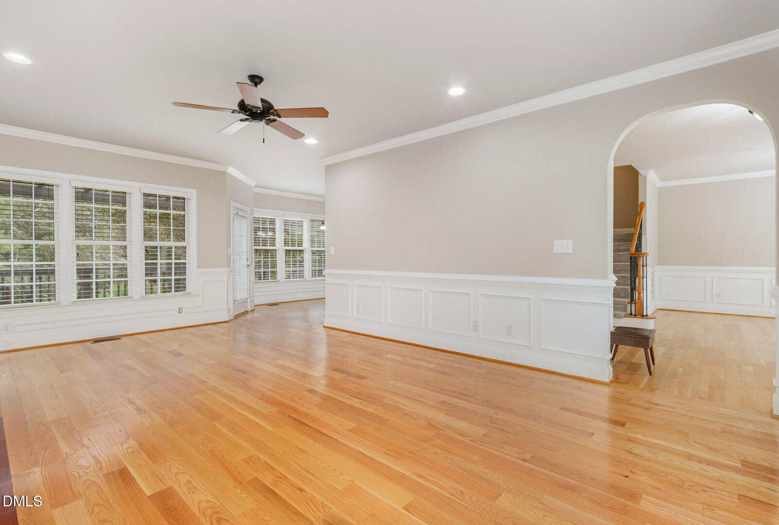 Spacious, empty living room with light hardwood floors, beige walls, white wainscoting, large windows, ceiling fan, and an arched doorway leading to a staircase.