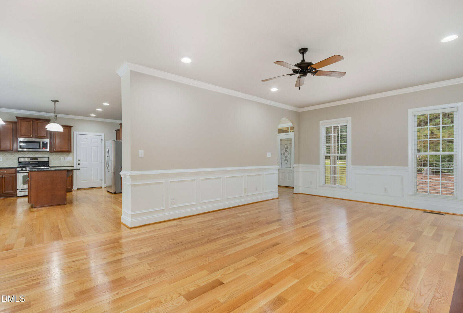 Open-concept living area with hardwood floors, beige walls, white trim, ceiling fan, large windows, and a view into a modern kitchen with dark wood cabinets and recessed lighting.