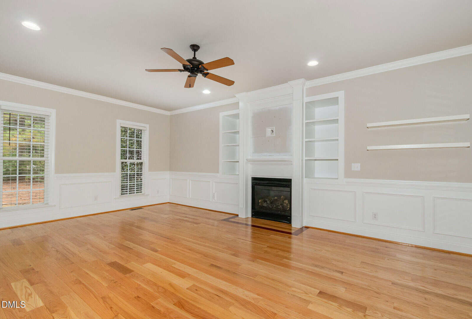 Bright living room with hardwood floors, white built-in shelves, a central fireplace, white walls with trim, two windows with blinds, ceiling fan, and recessed lighting.