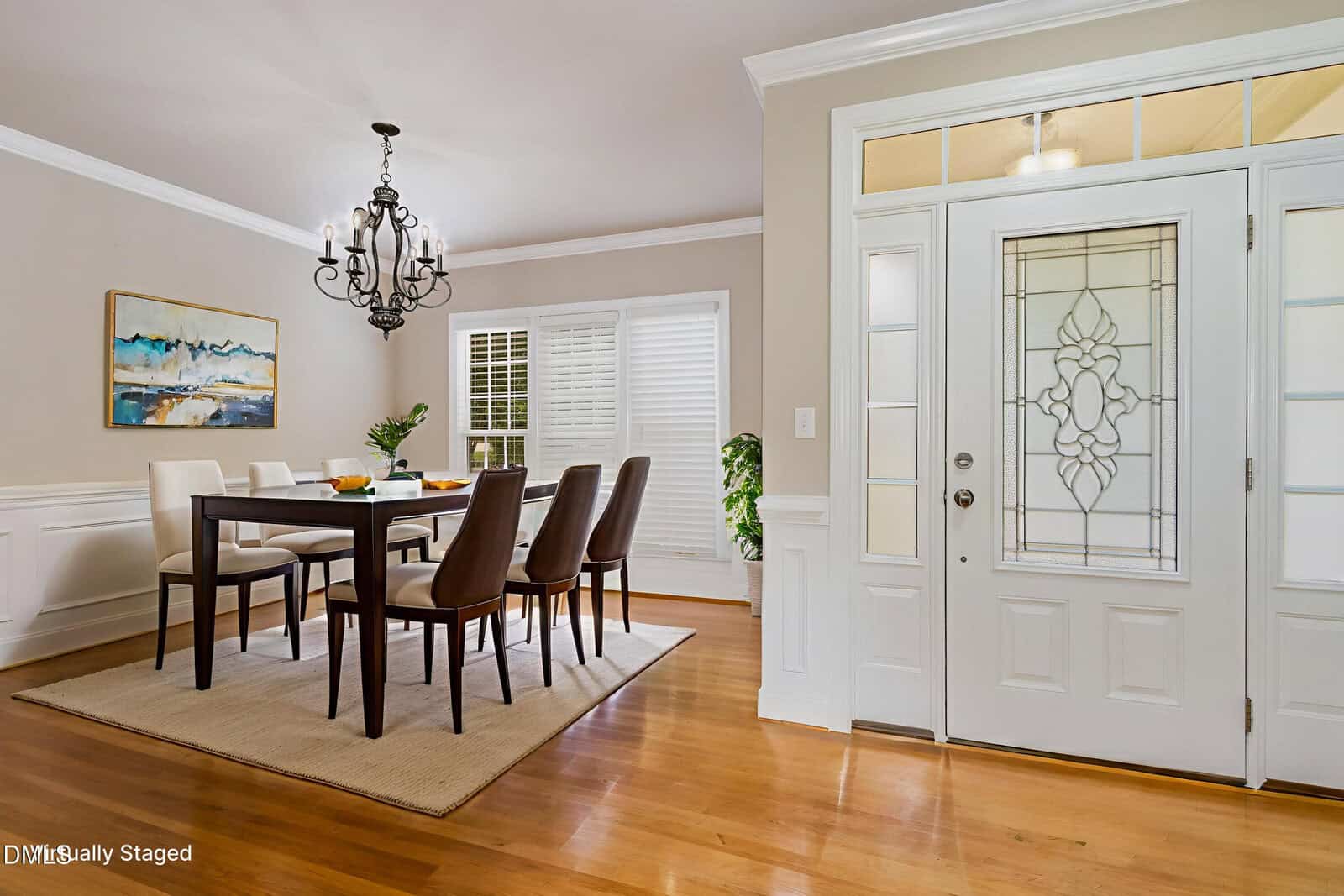 A bright, modern dining room with a rectangular table, six brown chairs, a chandelier, wall art, hardwood floors, and large windows with white shutters near a decorative glass front door.