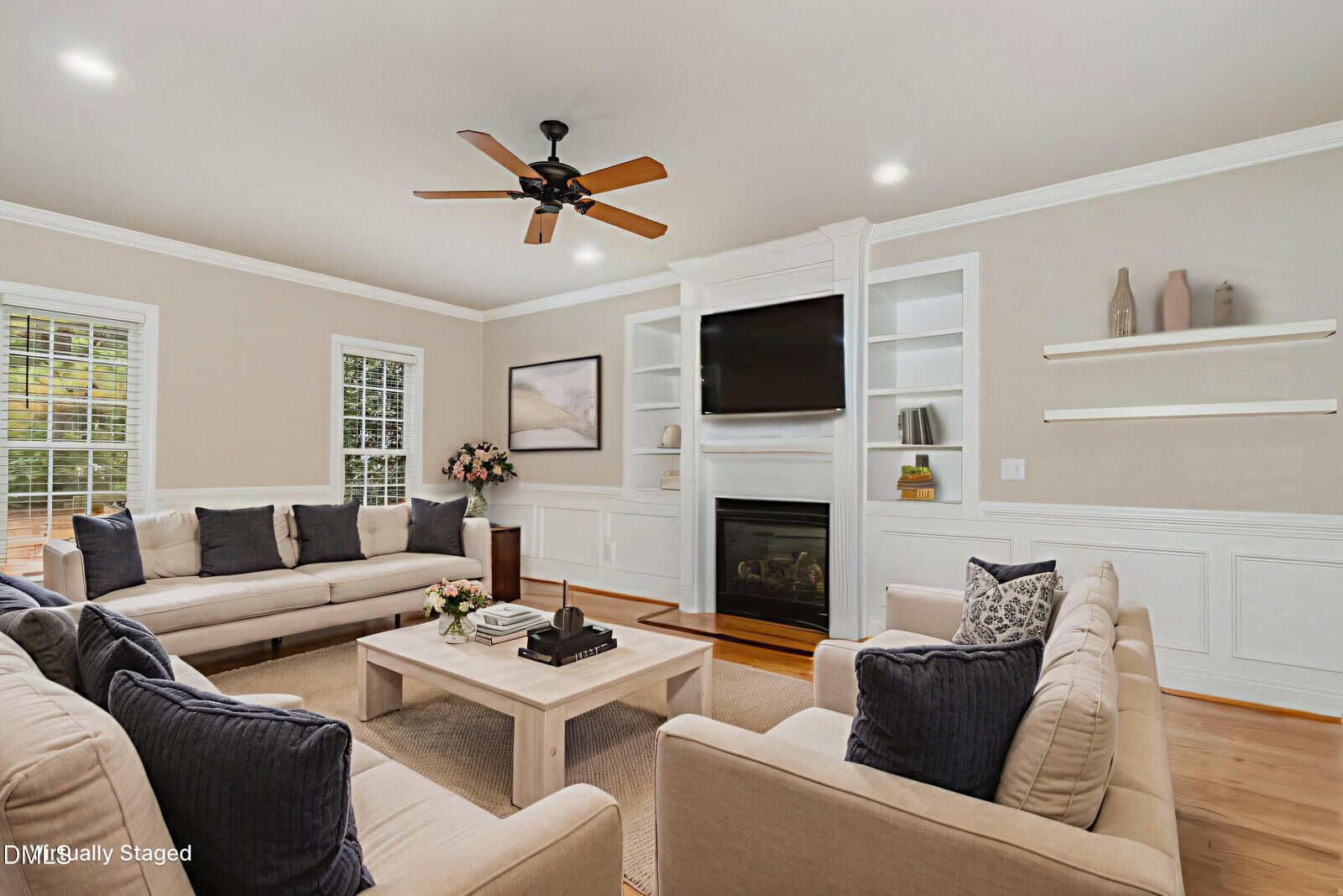 Bright living room with beige walls, two sofas, armchairs, a light wood coffee table, wall-mounted TV above a fireplace, ceiling fan, built-in shelves, large windows, and neutral decor accents.