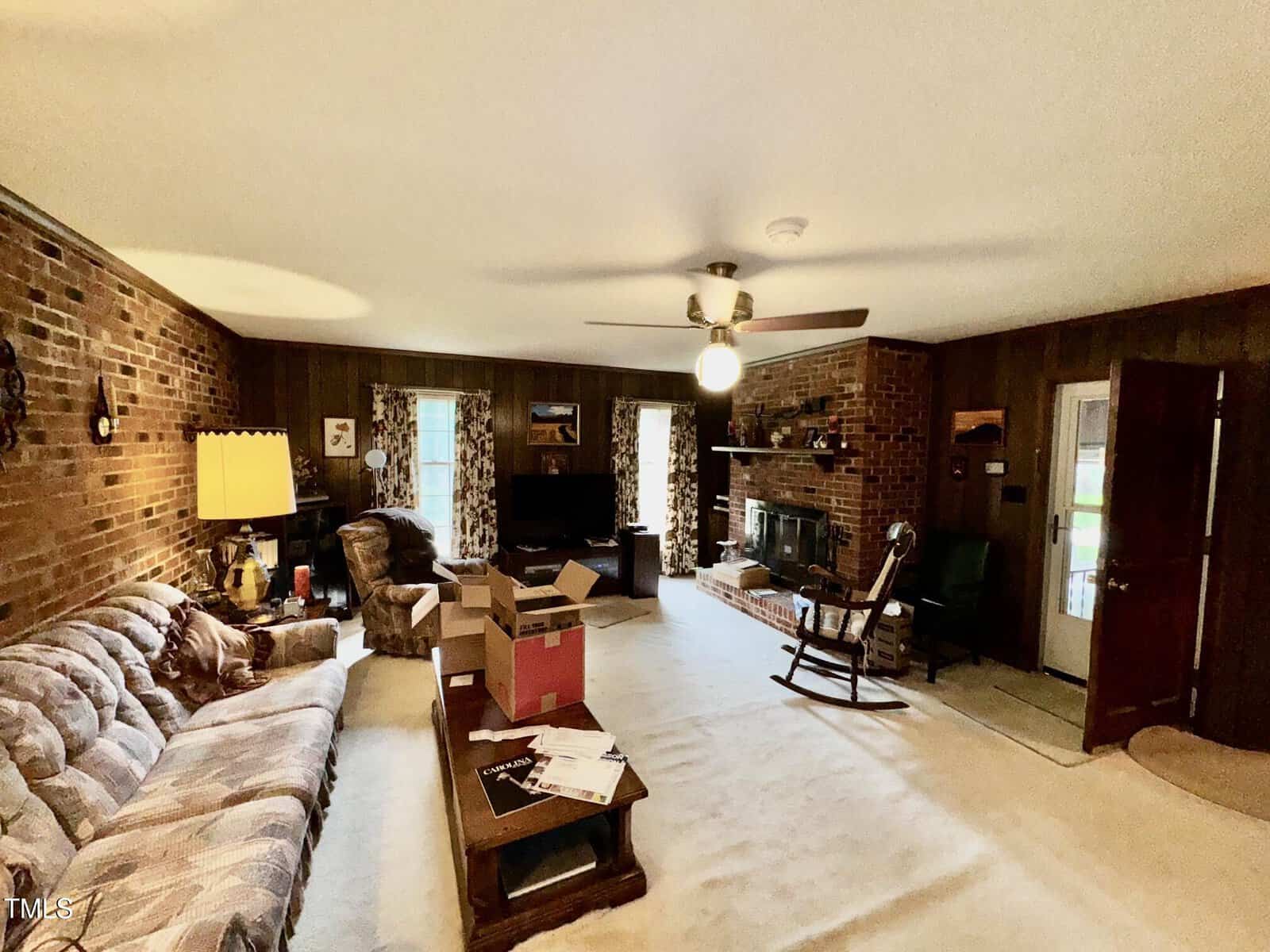 A spacious living room with brick and wood-paneled walls, a ceiling fan with a light, a sofa, armchairs, coffee table, boxes, a fireplace, TV, and a door leading outside. The room is carpeted and well-lit.