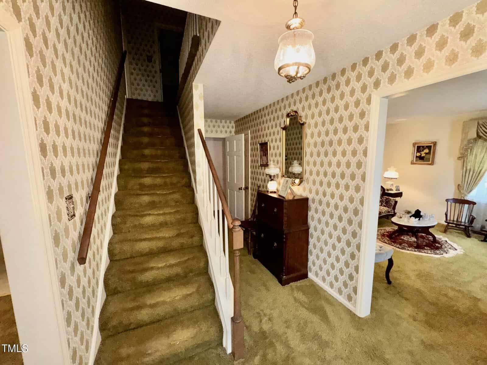 A hallway with patterned wallpaper, green carpeted stairs with a wooden handrail, a chandelier, a wooden dresser with a mirror, and a view into a living room with green carpet and classic furniture.