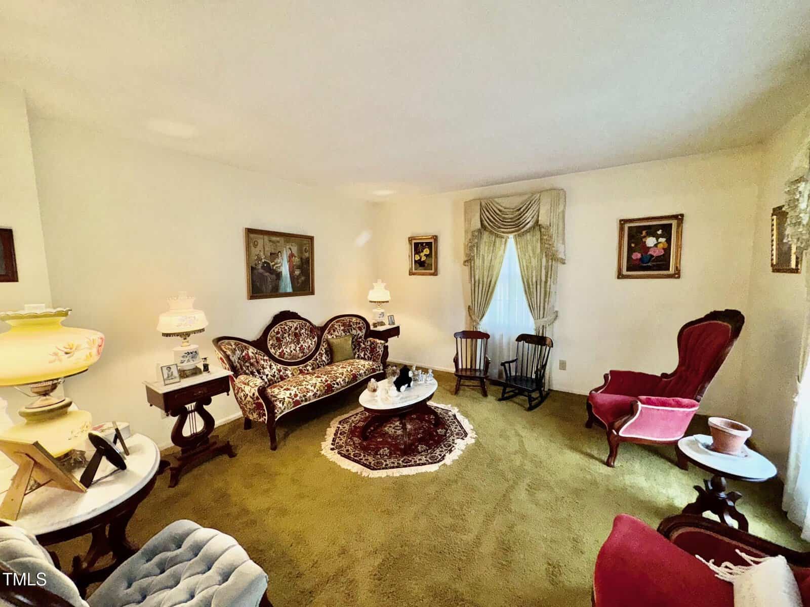 A living room with vintage furniture, including a patterned sofa, red armchair, wooden chairs, and ornate lamps. A small round table with a doily sits on a rug over green carpet. Framed art hangs on cream-colored walls.