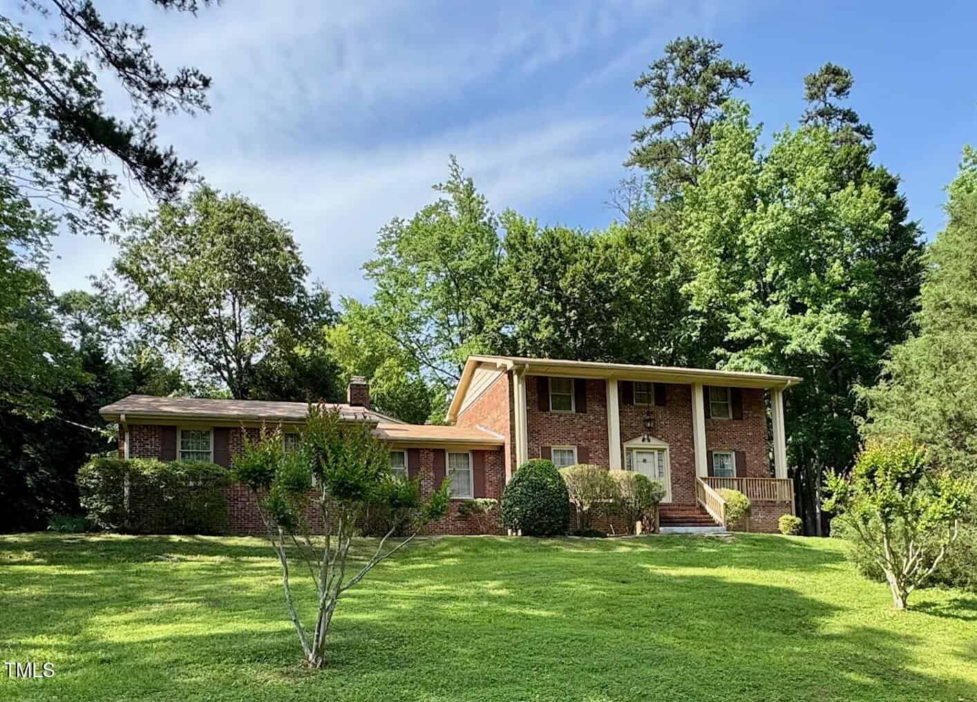 A two-story red brick house with white trim sits on a large, well-maintained lawn at 1818 Brookrun Road in Henderson. Mature trees and shrubs surround the home, which features a prominent front entrance with columns under a partly cloudy sky.