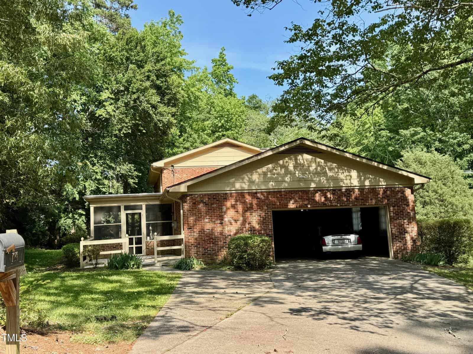 A brick house with a two-car garage, one door open showing a parked car inside. The home has a screened porch on the left and is surrounded by green trees and grass under a clear sky.