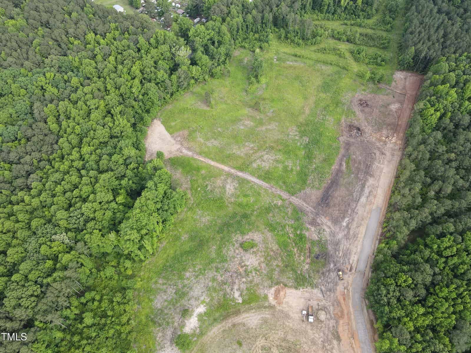 Aerial view of a partially cleared grassy area bordered by dense trees, with a dirt road and construction equipment visible along one edge. Some bare and disturbed patches are present among the greenery.