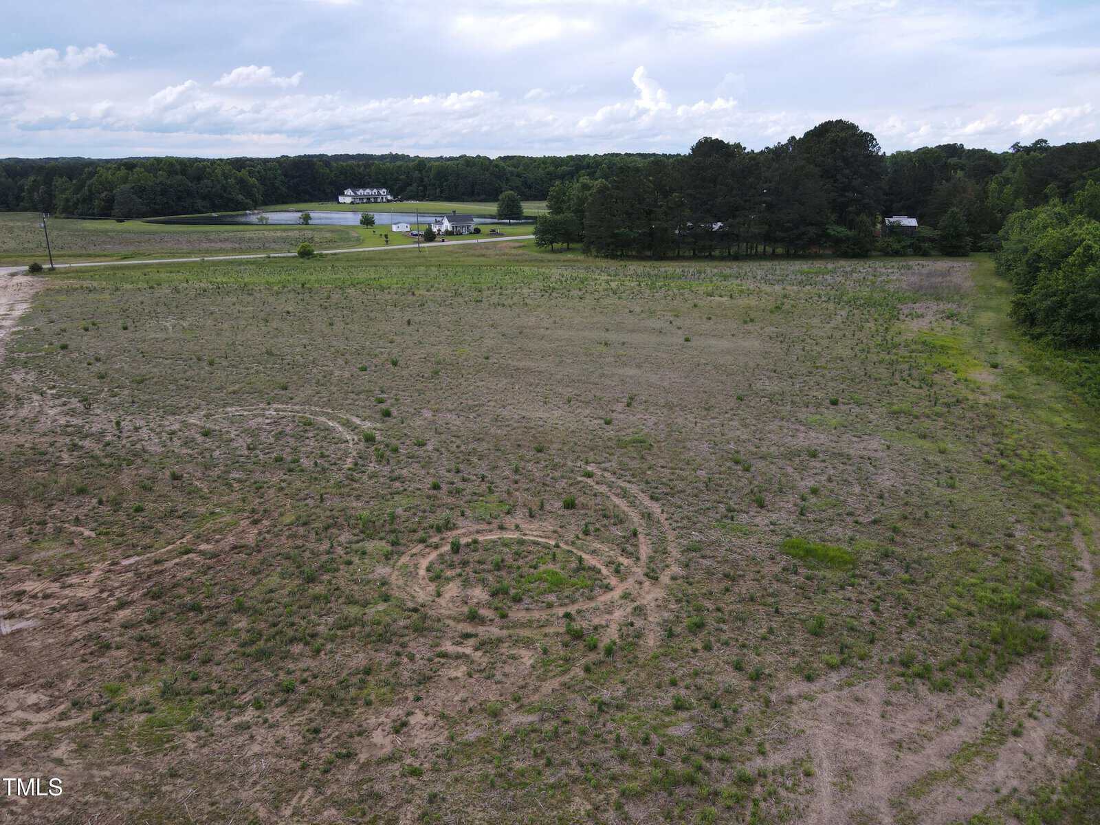 A mostly barren, open field with two visible circular marks in the dirt. Sparse grass and small plants cover parts of the ground. Trees line the right and background, with a pond, houses, and road in the distance under a cloudy sky.
