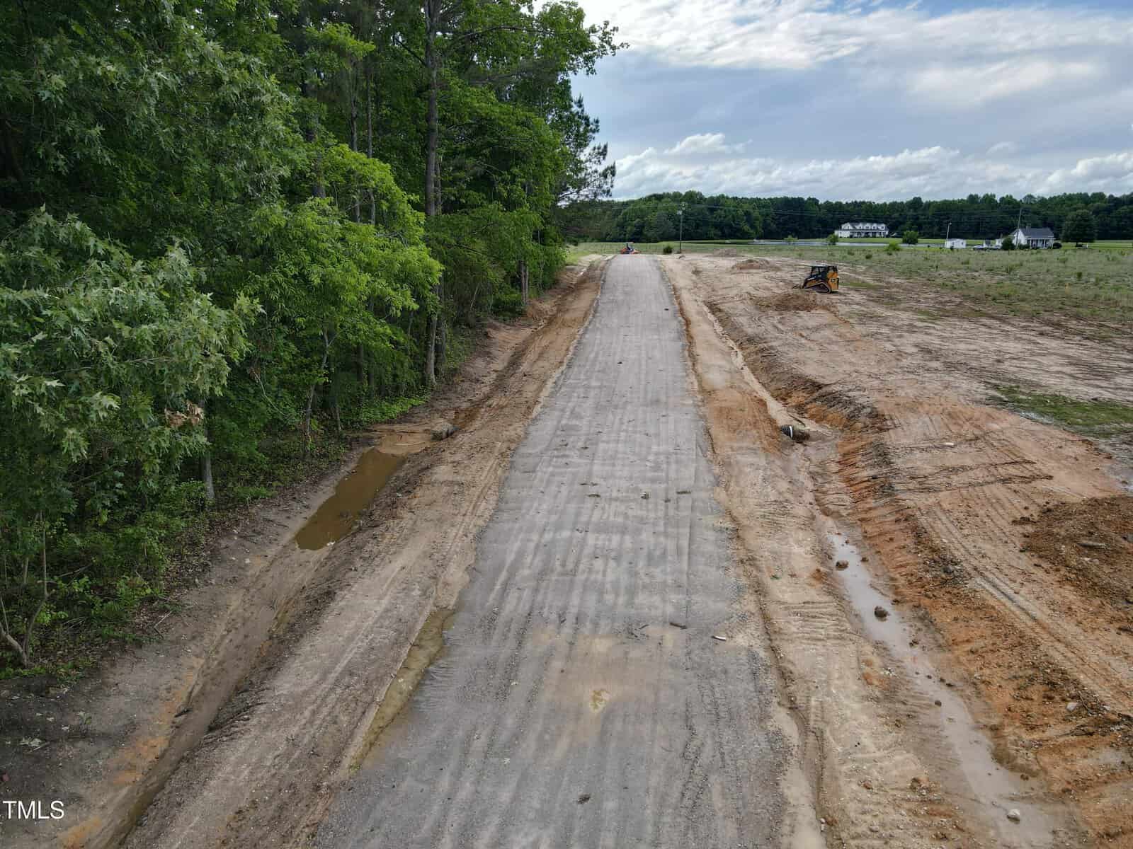 A newly paved road under construction runs through a rural area, bordered by trees on the left and cleared land on the right, with a bulldozer and some puddles visible.