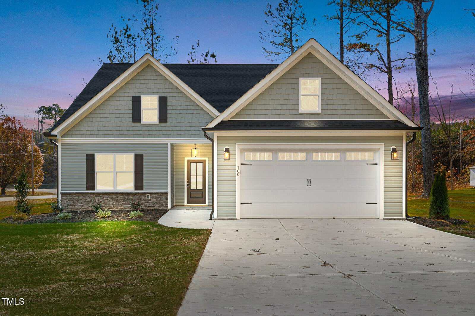 A modern, single-story house with gray siding, stone accents, a two-car garage, and warm lights glowing at dusk. The home sits on a spacious lot with a neat lawn and tall trees in the background.