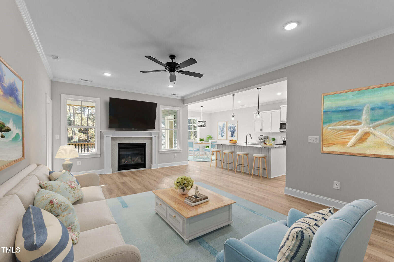 Bright, modern living room with neutral walls, a sofa, armchairs, and a coffee table. A fireplace under a large TV is against one wall, with an open view into a white kitchen and dining area. Coastal artwork decorates the space.