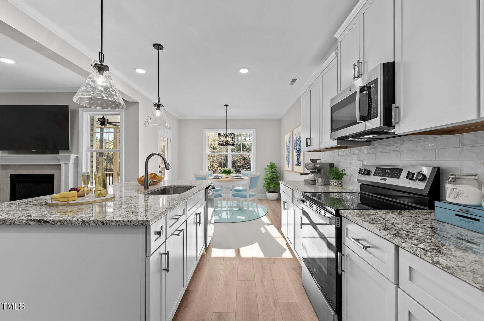 Modern kitchen with white cabinets, stainless steel appliances, granite countertops, and a central island. Sunlit dining area with a round glass table and blue chairs is visible through large windows in the background.