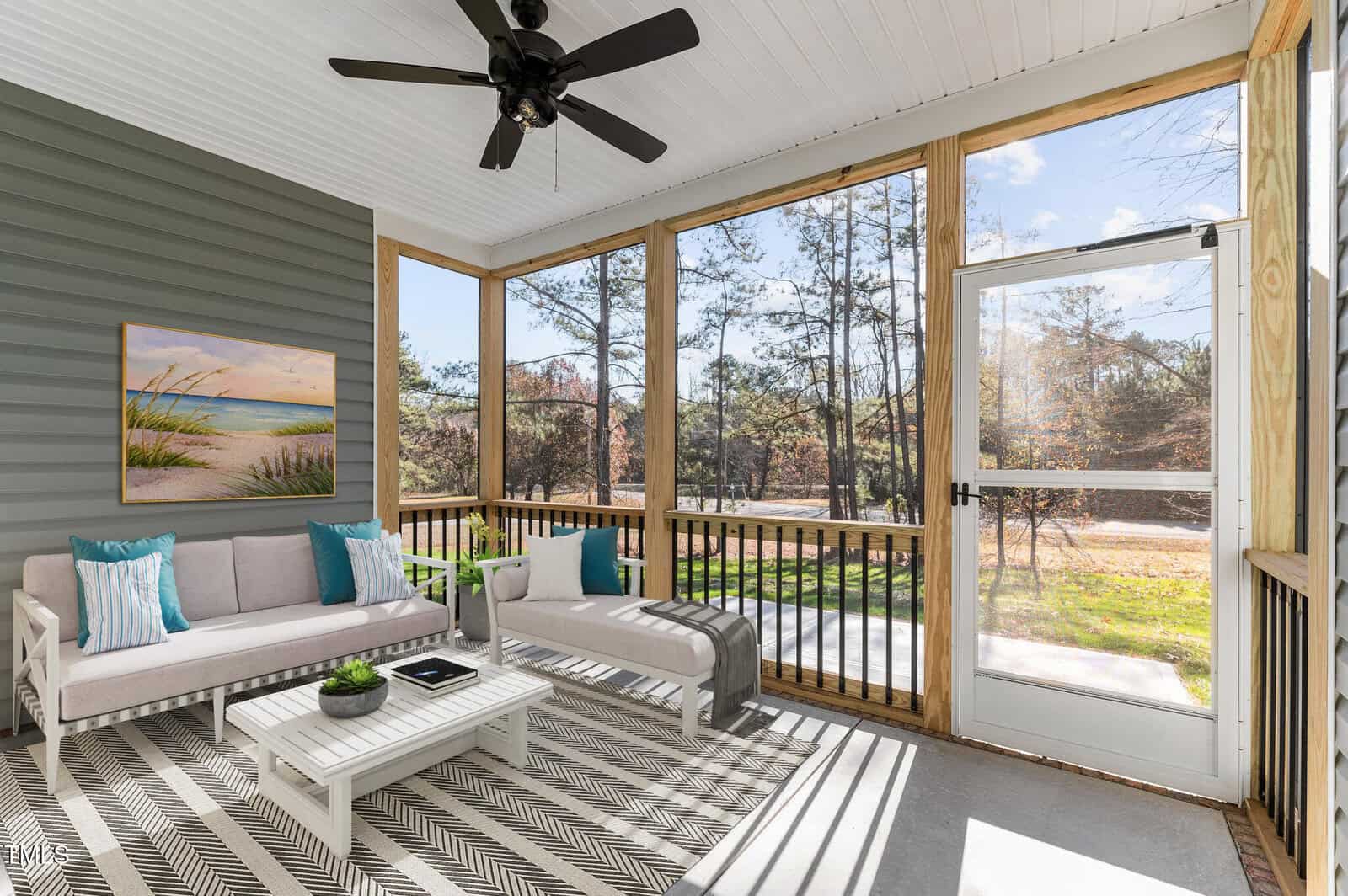 A screened-in porch with two light gray sofas, blue and white pillows, a small coffee table, a patterned rug, a ceiling fan, a landscape painting on the wall, and views of trees and grass through the screens.