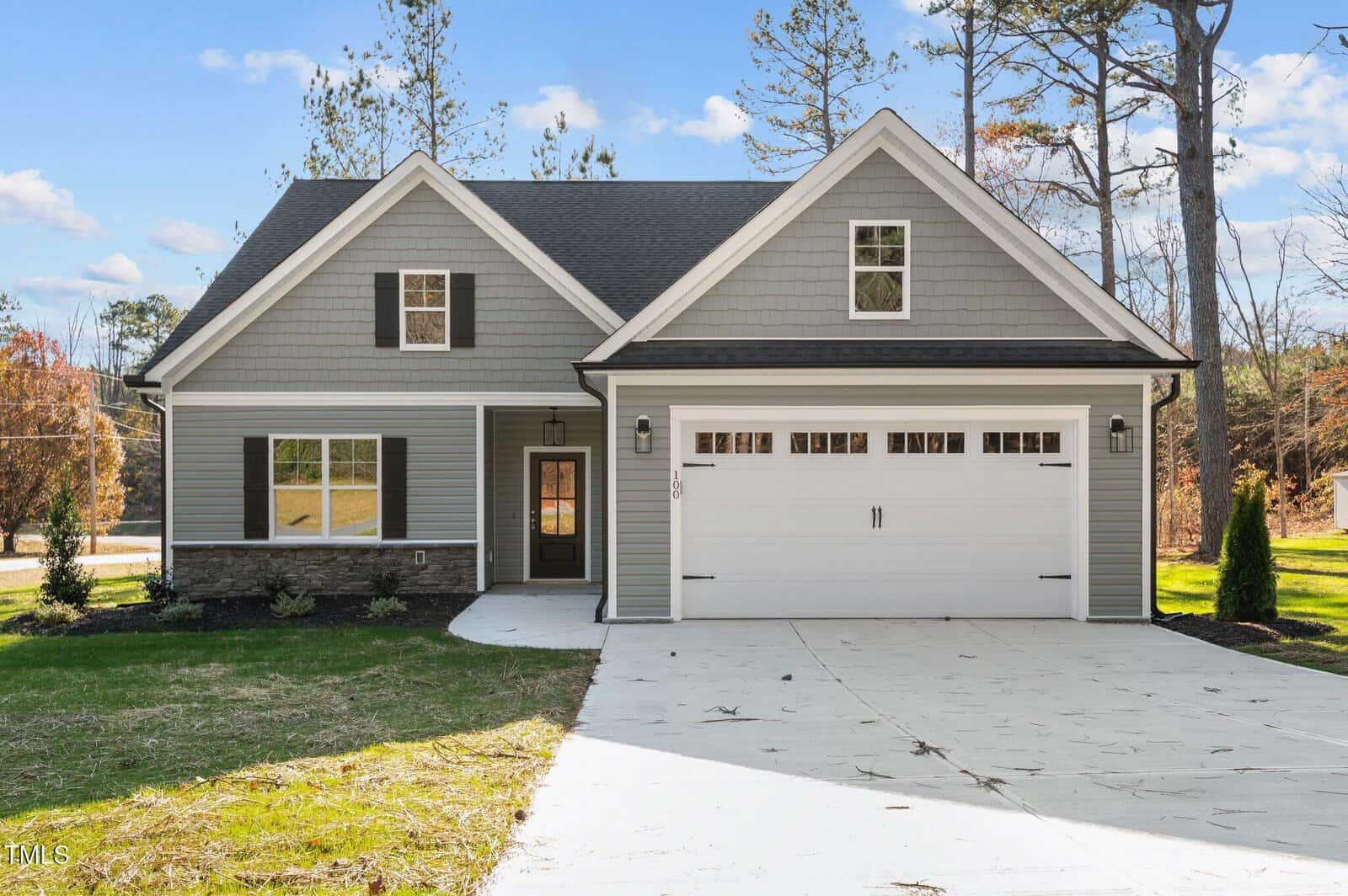 Single-family house with gray siding, white trim, and a double garage. Dark shutters frame the windows, and the home is surrounded by a neat lawn, young trees, and a driveway leading up to the garage.