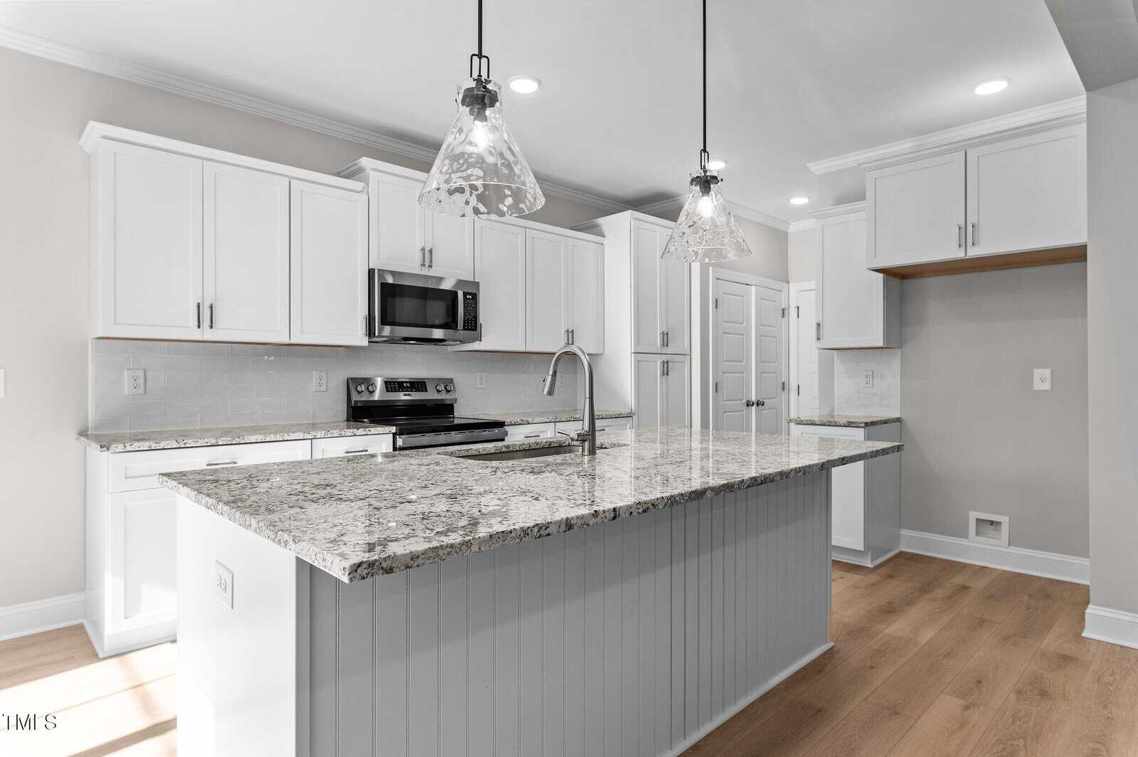 Modern kitchen with white cabinets, granite countertops, stainless steel appliances, an island with a sink, two pendant lights above the island, light wood flooring, and neutral-colored walls.