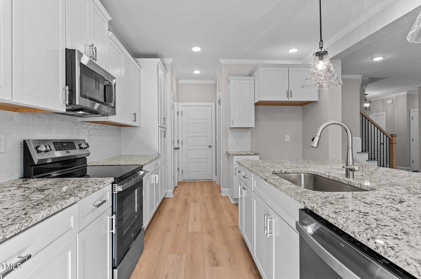 Modern kitchen with white cabinets, stainless steel appliances, granite countertops, a center island with a sink, hardwood floors, pendant lighting, and a door at the back wall. The space is clean and well-lit.