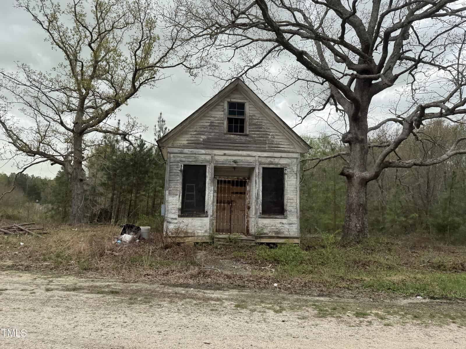 A small, old, white wooden house with boarded-up windows and a decaying exterior stands alone beside a dirt road. Overgrown grass and trees surround the abandoned structure under a cloudy sky.