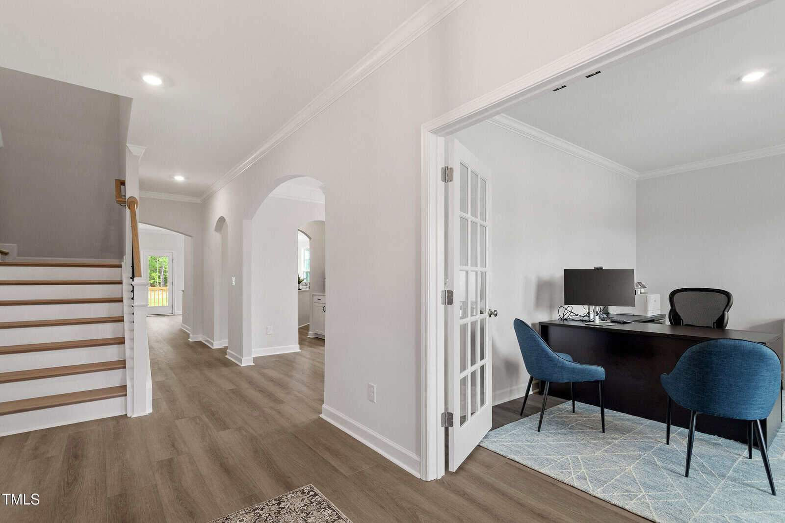 A modern home interior with light wood floors, white walls, a staircase on the left, an arched hallway, and a home office with a desk, computer, and two blue chairs on the right.