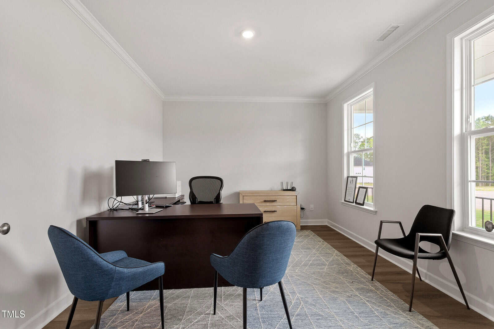 A modern office with a dark wood desk, office chair, computer monitor, two blue chairs, a black chair, a light wood cabinet, and large windows letting in natural light.