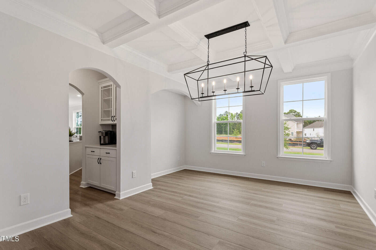 A bright, empty dining room with light wood flooring, coffered ceiling, modern rectangular chandelier, two large windows, and an arched doorway leading to a kitchen area.