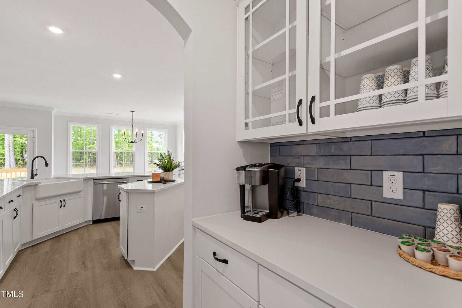 A modern kitchen with white cabinets, a farmhouse sink, stainless appliances, and a nook featuring a coffee maker, mugs, and K-cups. The backsplash is blue tile, and large windows provide natural light.