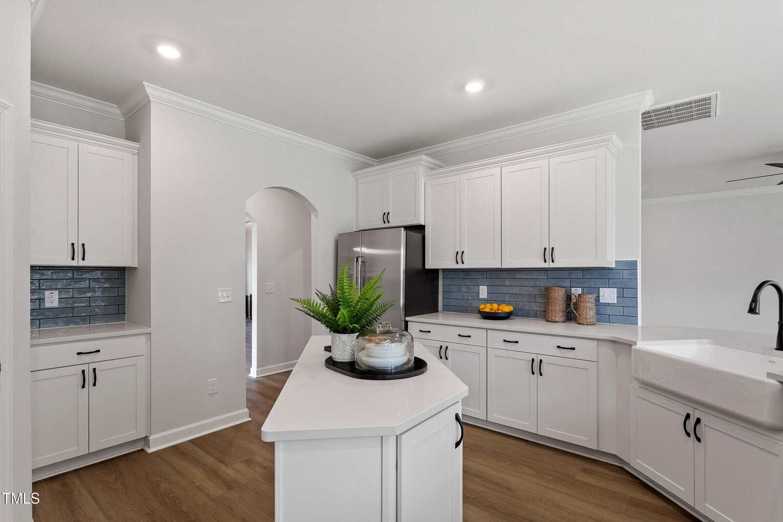 Modern kitchen with white cabinets, blue subway tile backsplash, stainless steel appliances, farmhouse sink, wood floors, and a small island with a plant centerpiece. Bowl of lemons and two containers are on the counter.