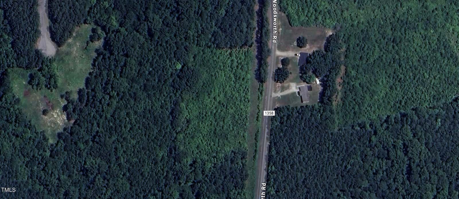 Aerial view of a rural wooded area with a road running vertically through the center. A few buildings and cleared spaces are visible on the right side of the road, surrounded by dense trees.