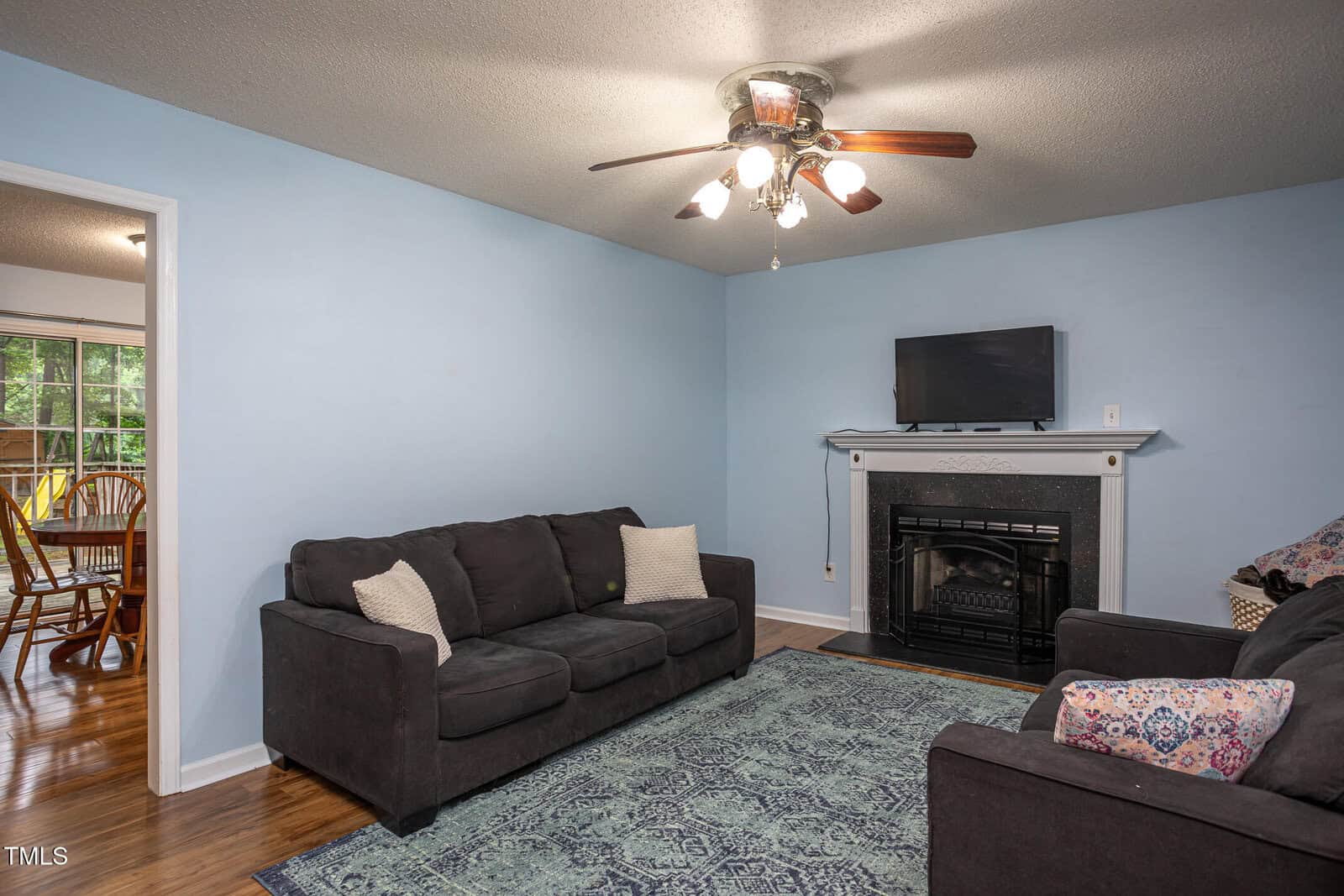 A living room with light blue walls, a ceiling fan with lights, a dark sofa and loveseat, a patterned rug, and a fireplace with a TV on the mantel. An adjacent dining area with wooden furniture is visible through an open doorway.