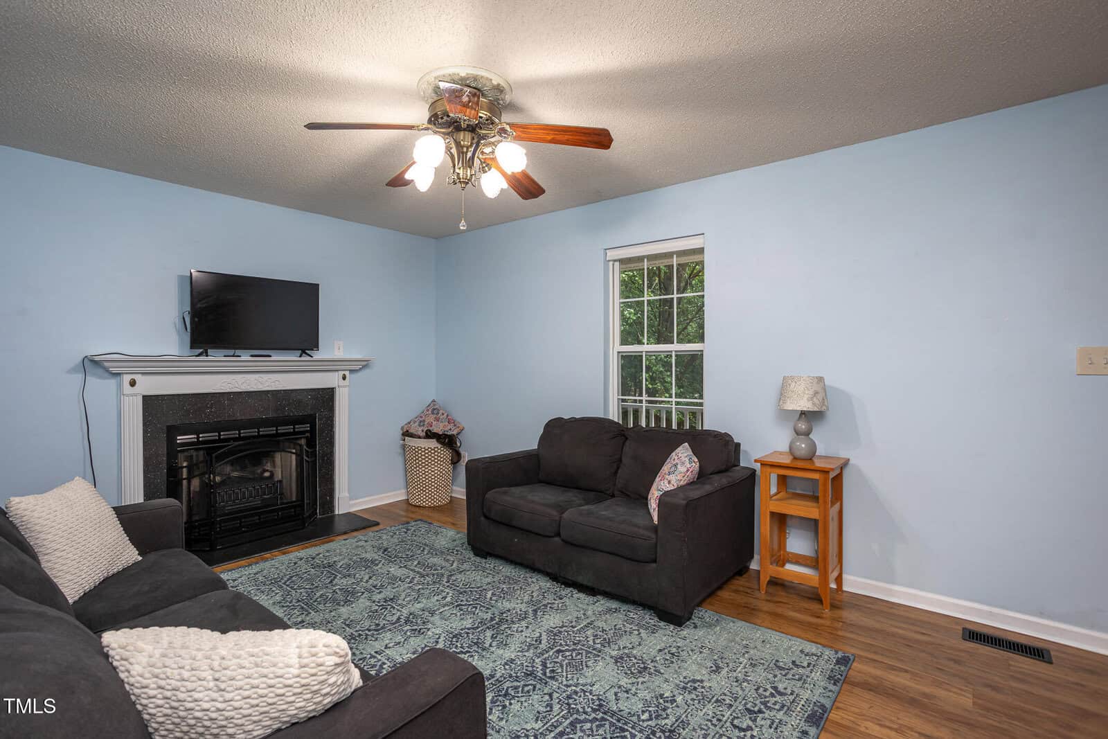 A cozy living room with light blue walls, a ceiling fan with lights, two dark couches with pillows, a patterned rug, a fireplace with a TV above it, a basket, and a side table with a lamp next to a window.