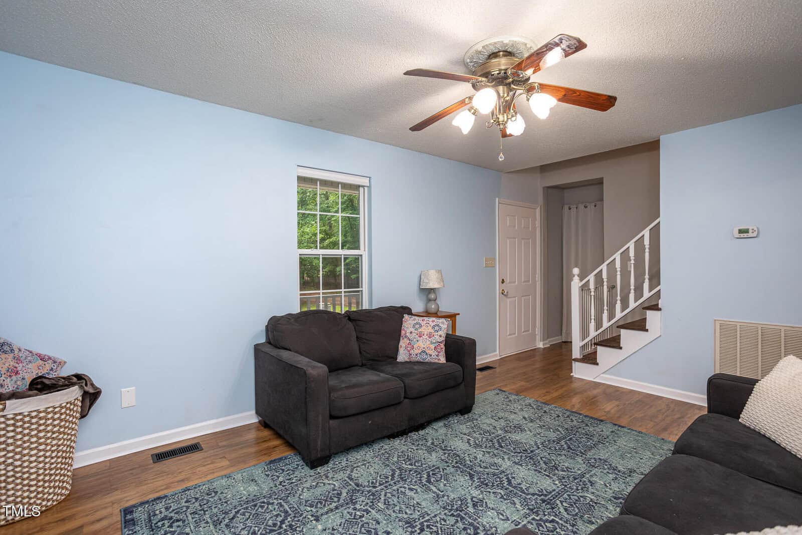Living room with light blue walls, dark sofas, patterned rug, a laundry basket, wooden floors, a ceiling fan with lights, a window, a lamp on a small table, and stairs leading to an upper level.