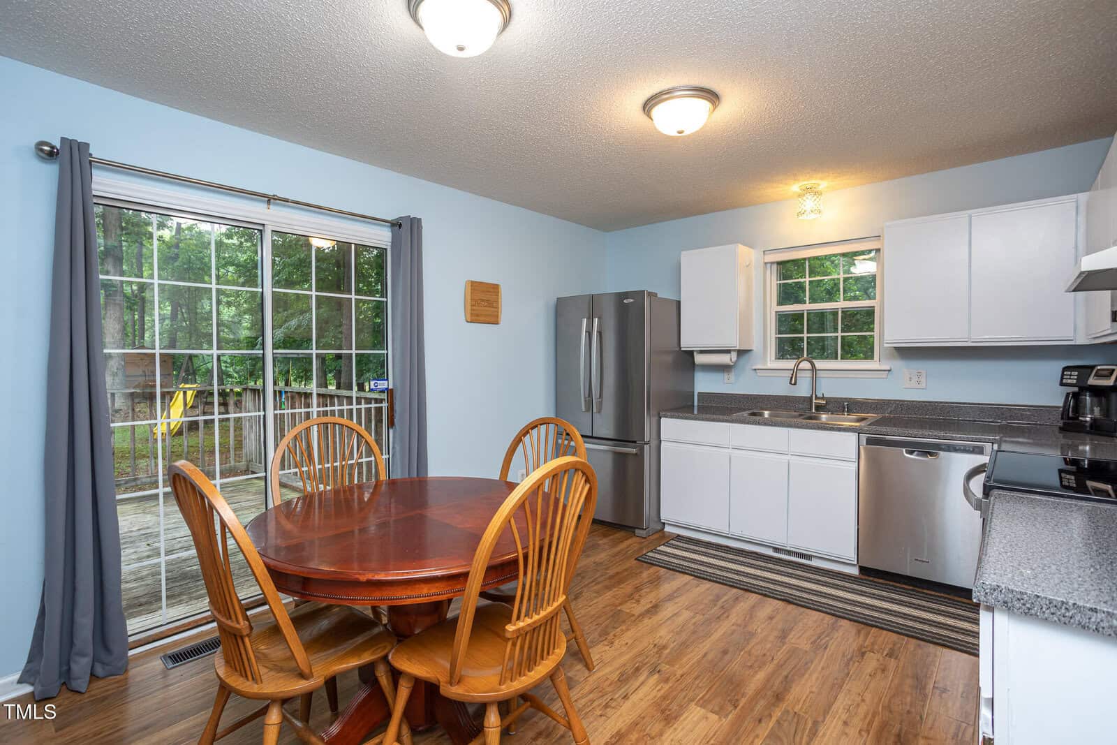 A kitchen with white cabinets, stainless steel appliances, wood flooring, and a round wooden dining table with four chairs. Sliding glass doors with gray curtains open to a deck and a view of trees outside.