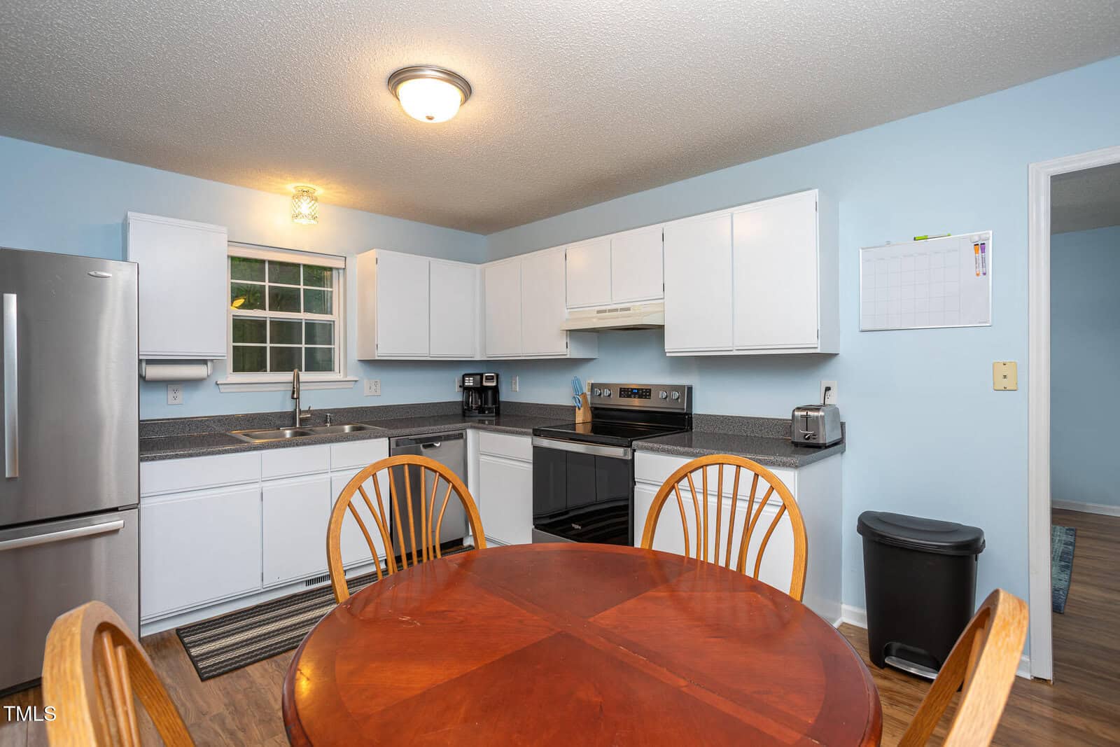 A kitchen with white cabinets, stainless steel fridge, stove, dishwasher, and a round wooden dining table with four chairs. Light blue walls, a wall calendar, trash can, and a window above the sink are visible.