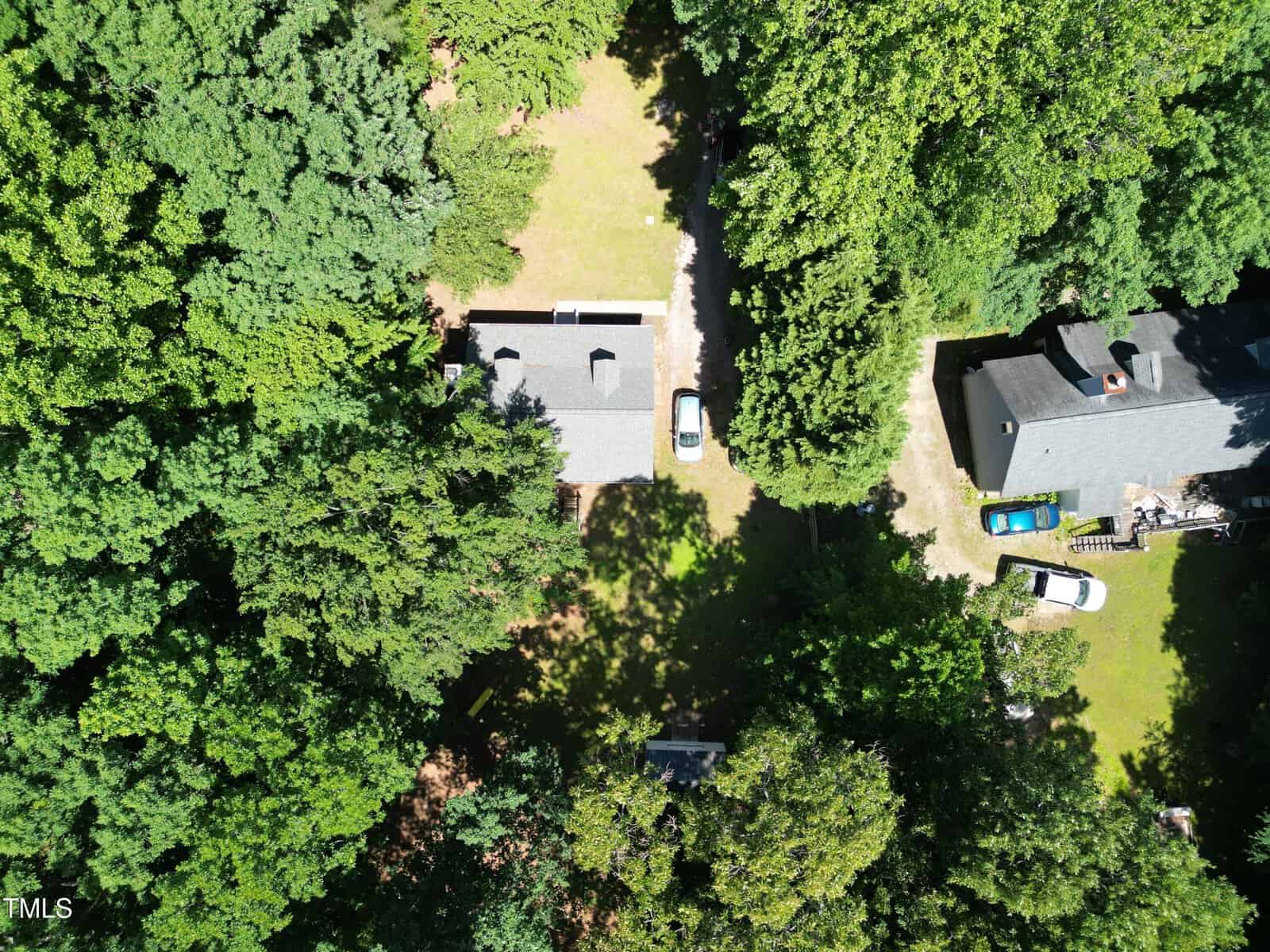 Aerial view of two houses surrounded by dense trees, with cars parked on driveways and grassy yards in between. Sunlight creates patches of light and shadow throughout the green landscape.