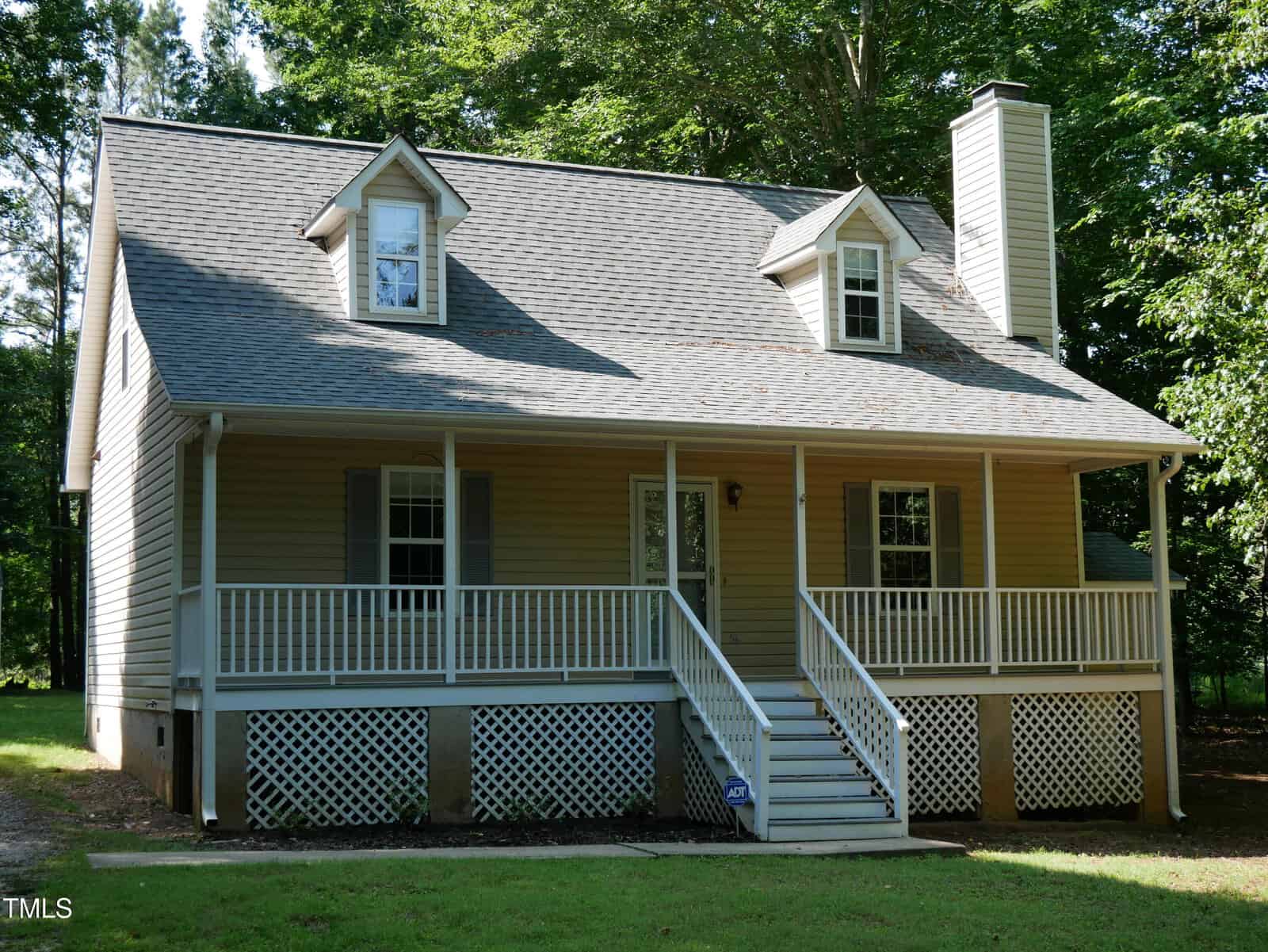 A beige two-story house with a large covered front porch, white railings, two dormer windows, a brick chimney, and surrounded by green trees under bright sunlight.