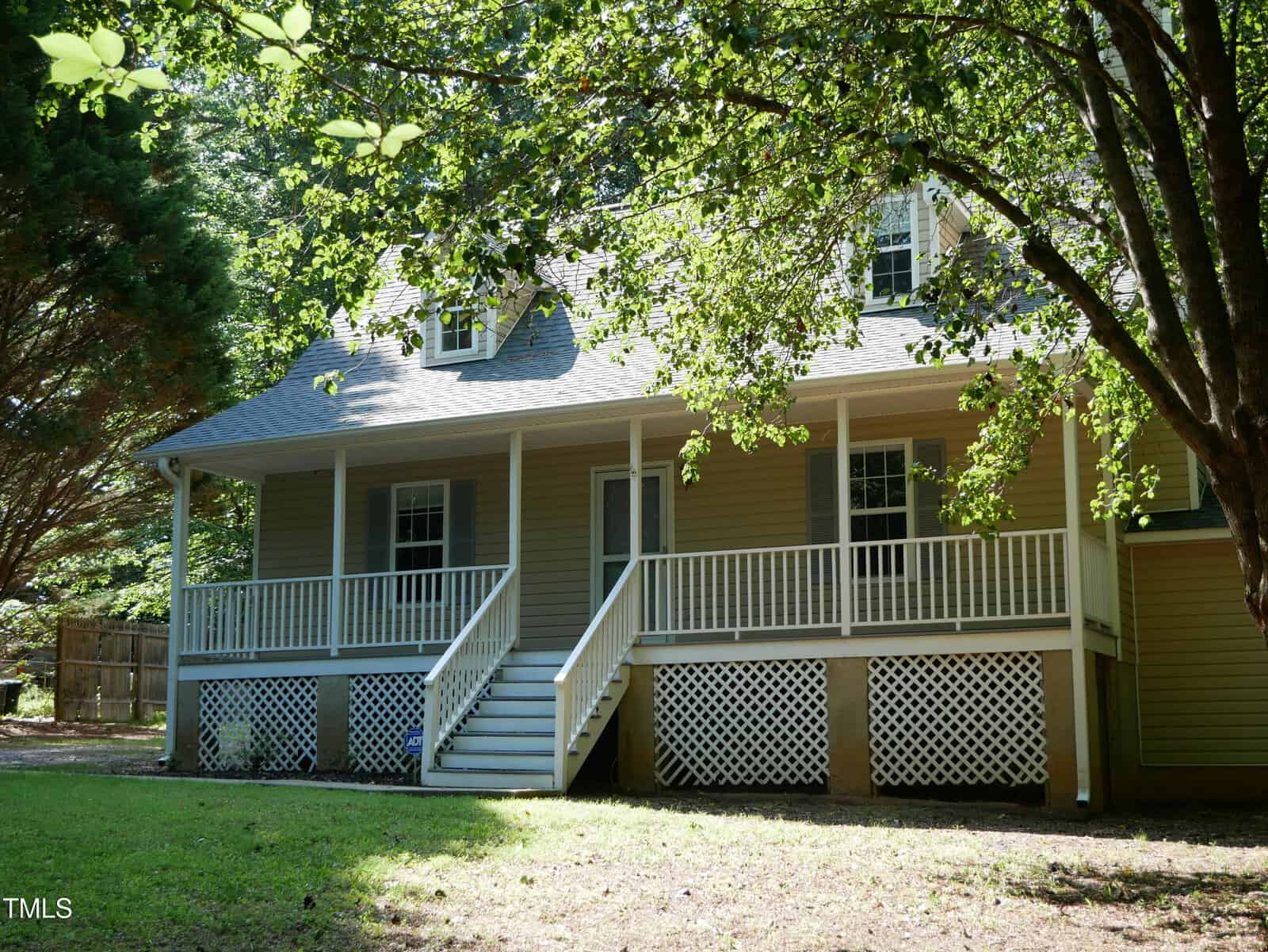 A beige house with a covered front porch, white railings, lattice panels, and steps leading up to the entrance. The yard is shaded by large green trees, and sunlight filters through the leaves.