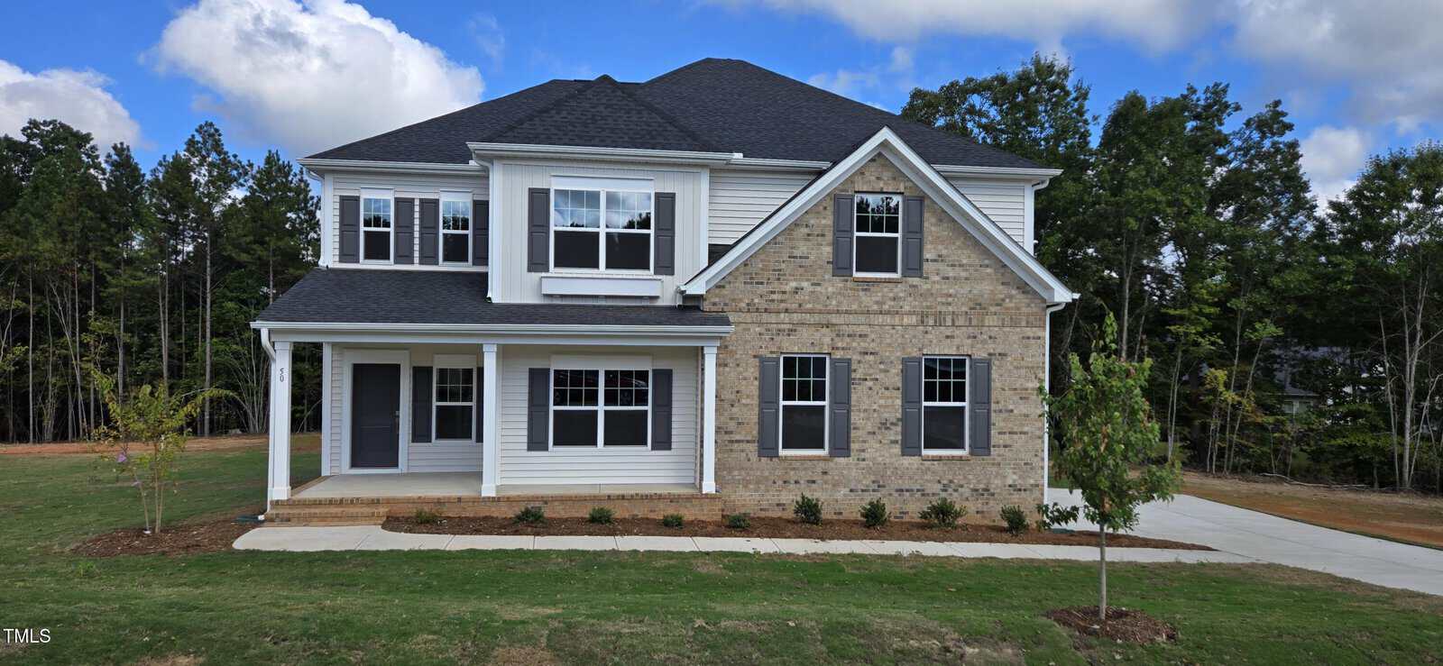 Two-story suburban house with a mix of brick and white siding exterior, black roof, multiple windows, covered front porch, driveway, small trees, and landscaped yard; surrounded by trees under a partly cloudy sky.