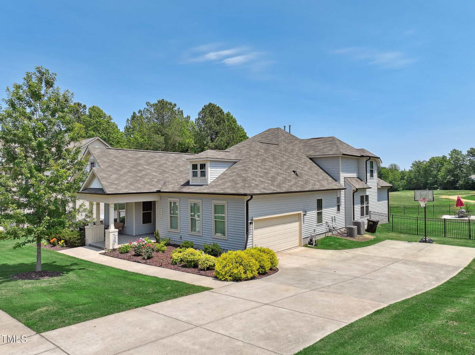 A two-story suburban house with light-colored siding, a covered porch, and an attached two-car garage. The driveway leads to a basketball hoop, and the yard is landscaped with shrubs and green grass. Trees surround the property.