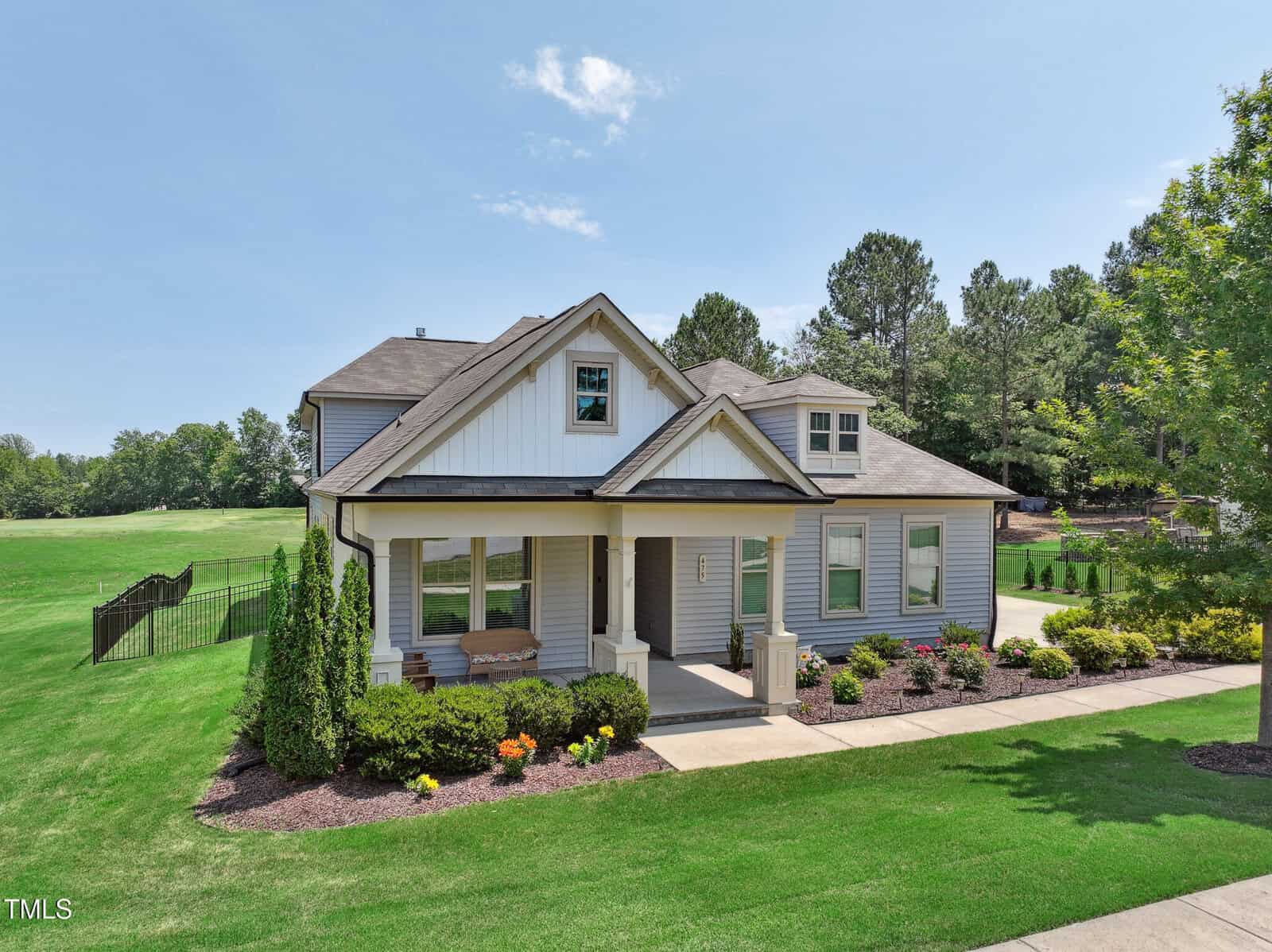 A modern two-story house with gray siding, white trim, and a covered front porch. The home is surrounded by green lawn, colorful flowers, and trees, with a driveway and a golf course in the background under a clear sky.
