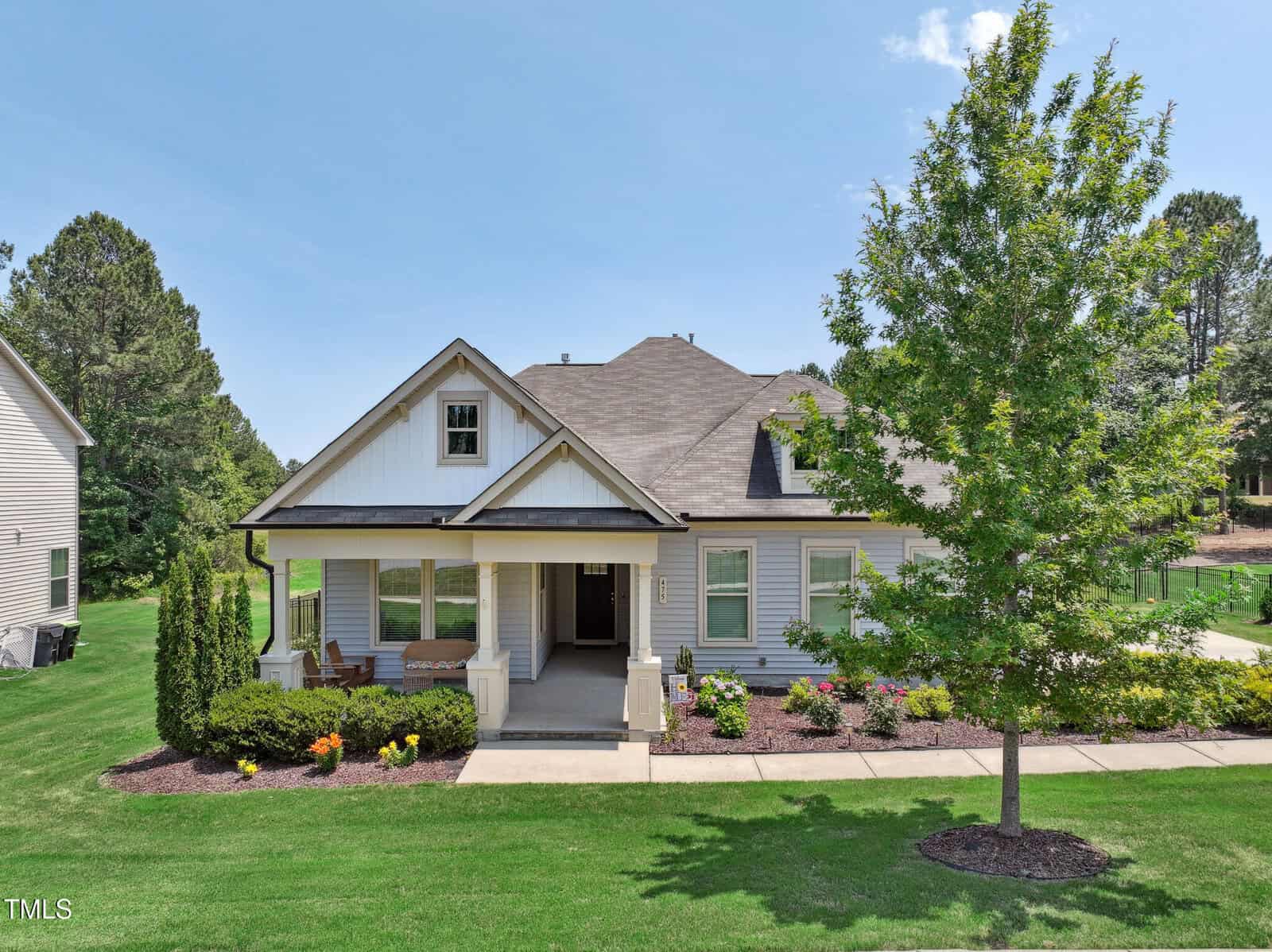 Single-story house with a front porch, white exterior, and gabled roof, surrounded by a well-kept lawn, shrubs, colorful flowers, and a large leafy tree in the front yard. Bright, sunny day with a clear blue sky.