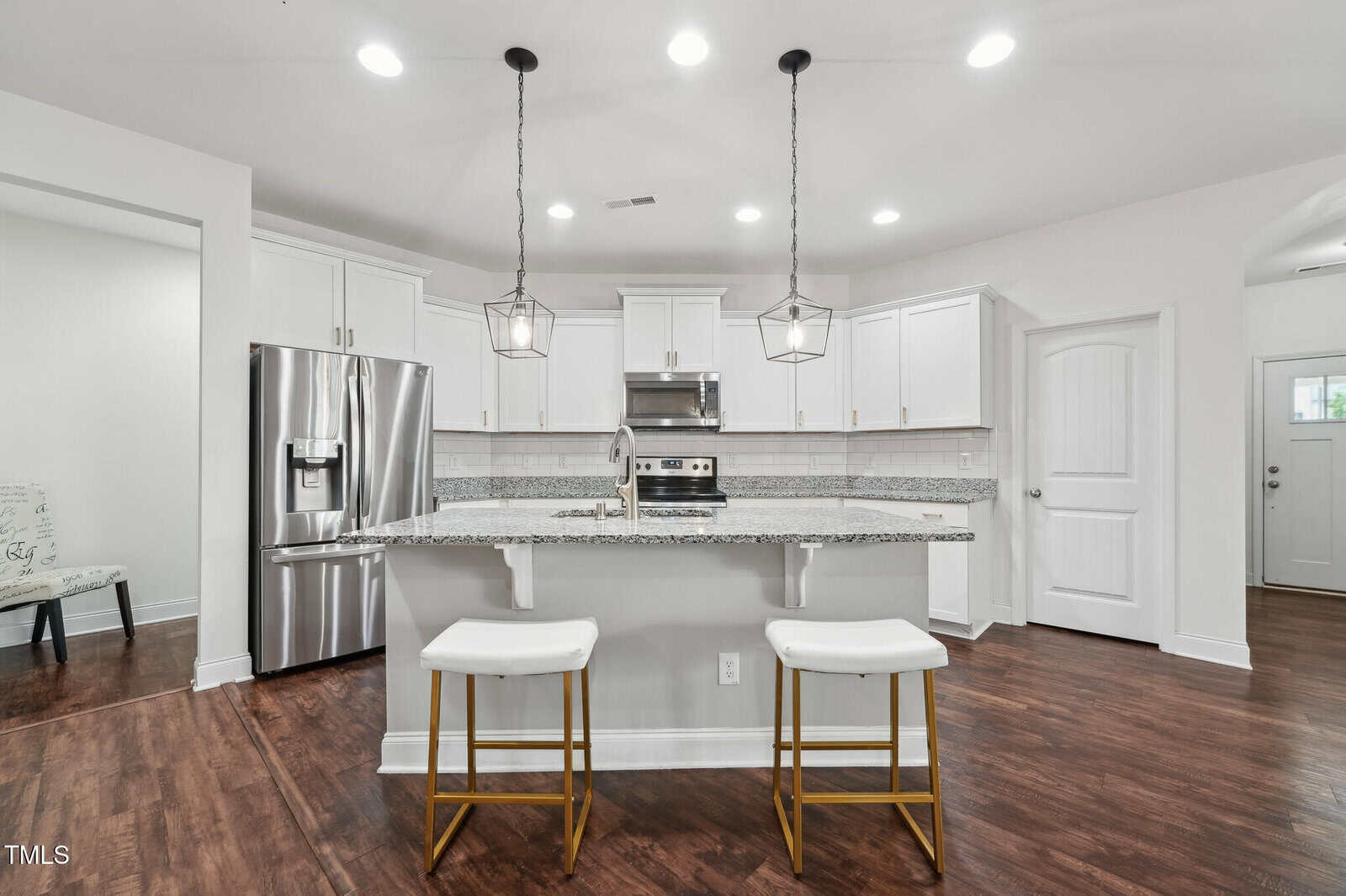 Modern kitchen with white cabinets, stainless steel appliances, granite island with two gold-accented white stools, pendant lights, wood flooring, and recessed ceiling lights. A door and hallway entry are visible in the background.
