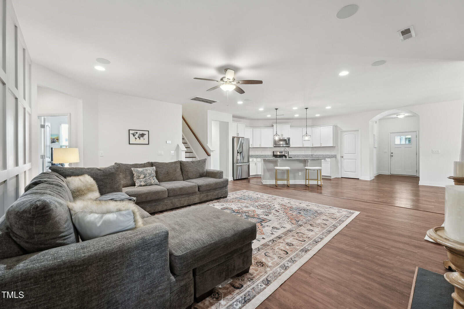 Open-concept living room and kitchen with hardwood floors, gray sectional sofa, area rug, ceiling fan, stainless steel appliances, white cabinetry, island seating, and a staircase leading up. Room is well-lit and spacious.