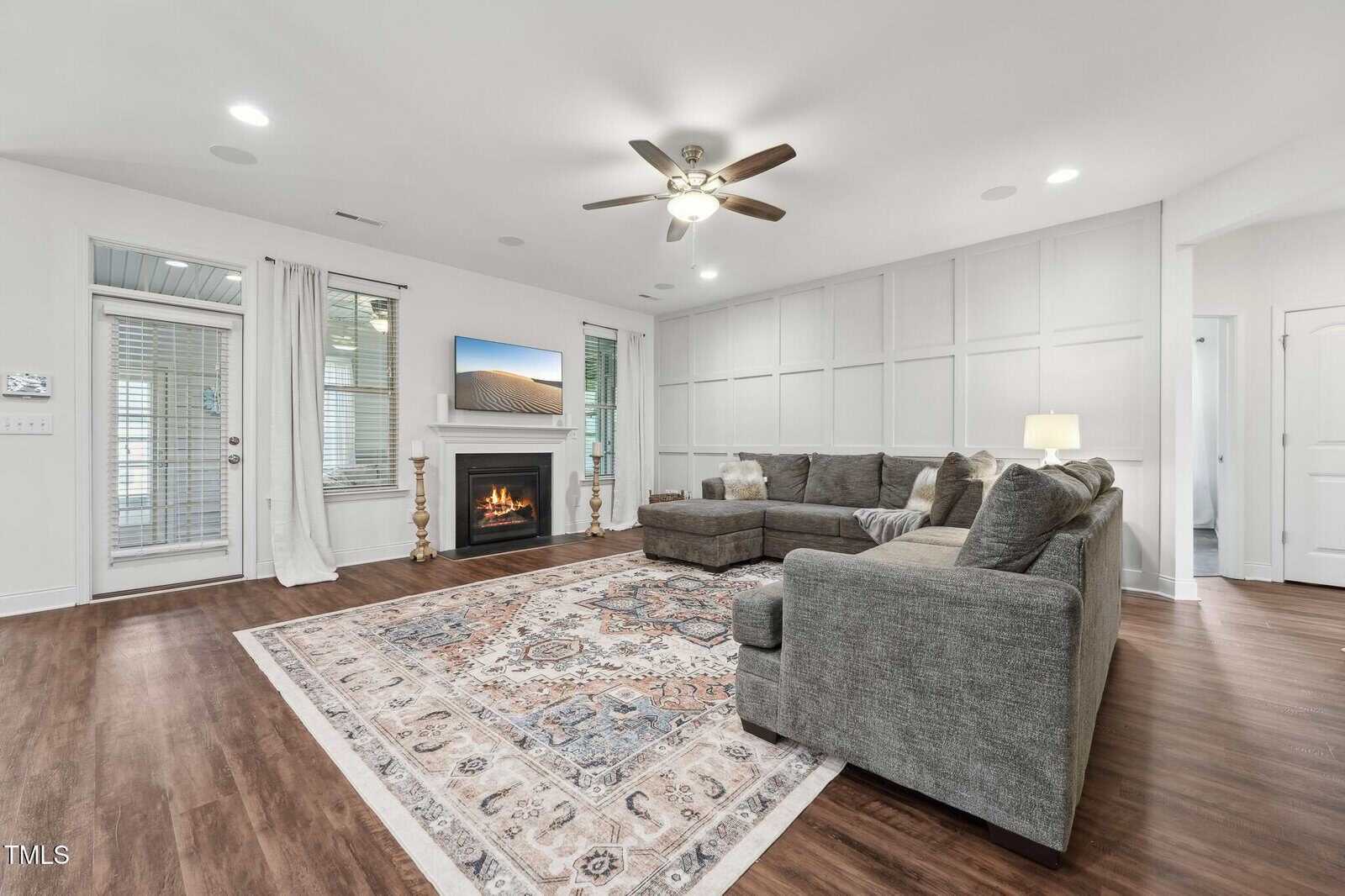 Spacious living room with wood floors, a large gray sectional sofa, patterned area rug, fireplace with a TV above, ceiling fan, and white paneled walls. French doors and windows let in natural light.