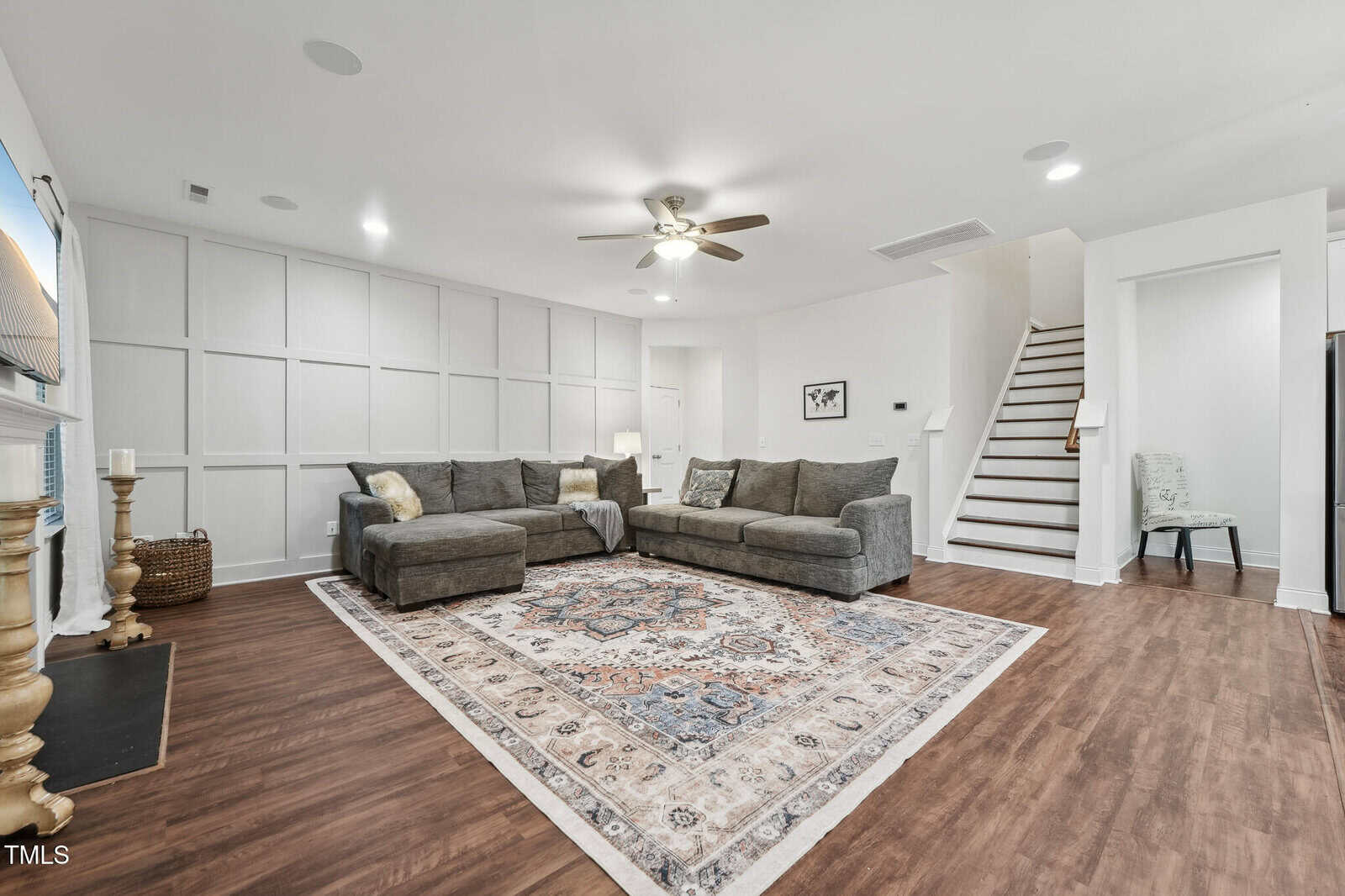 A spacious living room with wood floors, a large gray sectional sofa, an ornate area rug, white paneled walls, a ceiling fan, and a staircase leading upward. A chair is placed near the stairs.