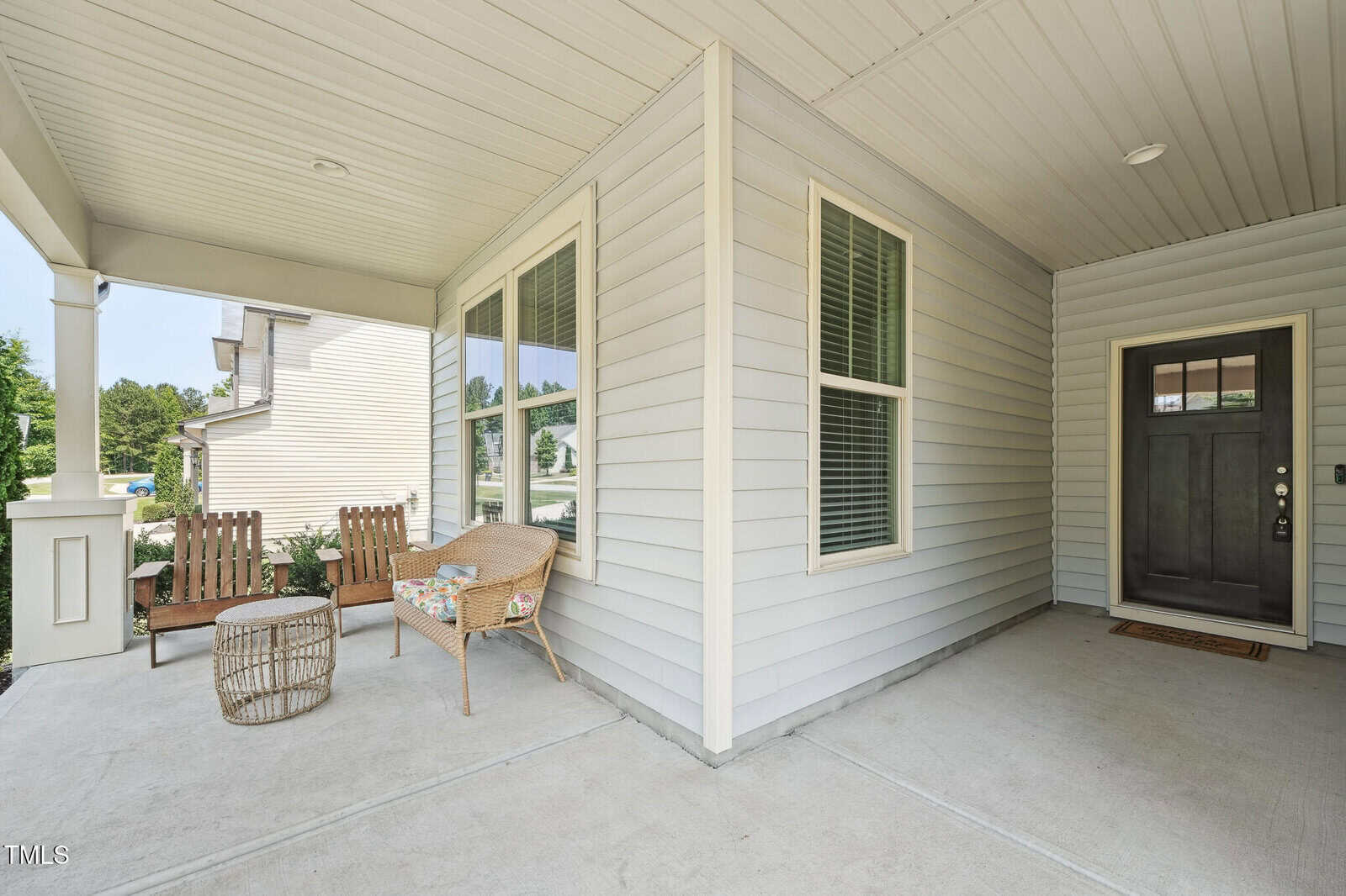 Covered front porch with light gray siding, a black door, two wooden chairs, a wicker chair with a cushion, a small round table, and potted plants; windows with white shutters and concrete flooring.