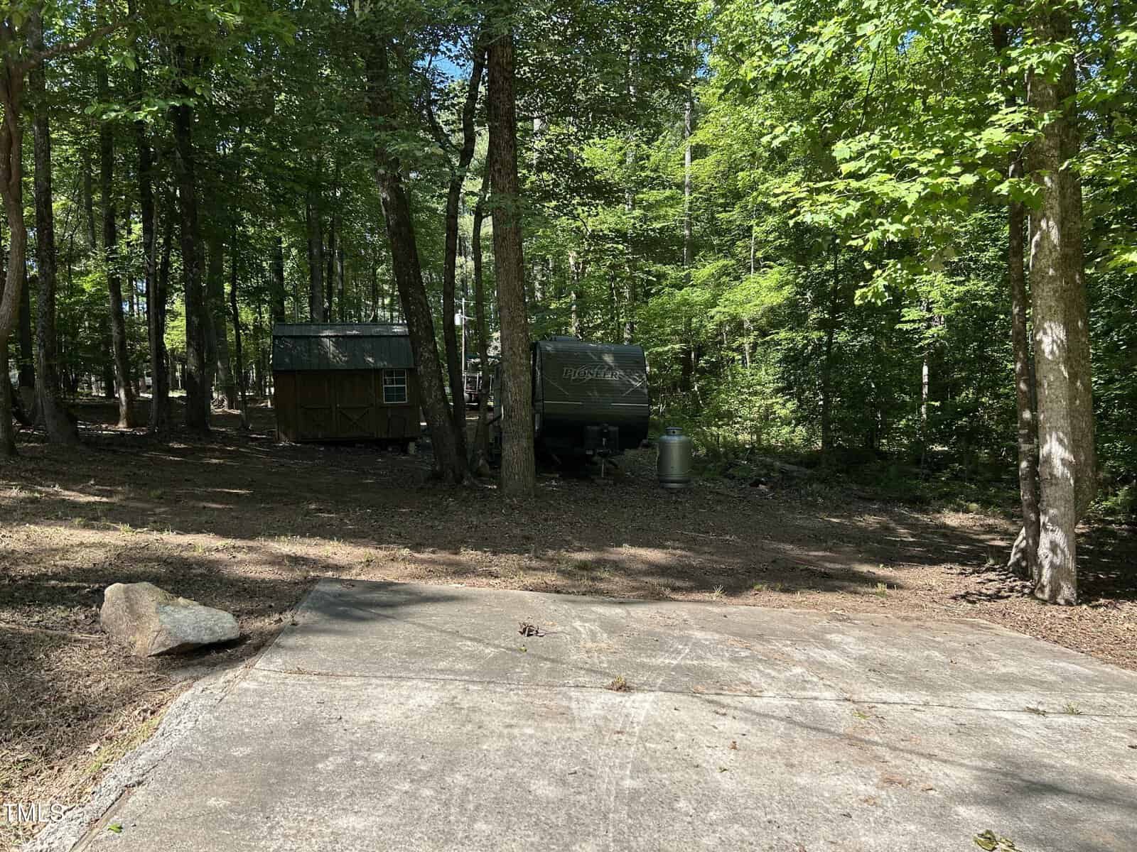 A small wooden shed and a camper trailer are nestled among tall trees in a wooded area, with a concrete pad in the foreground under dappled sunlight.