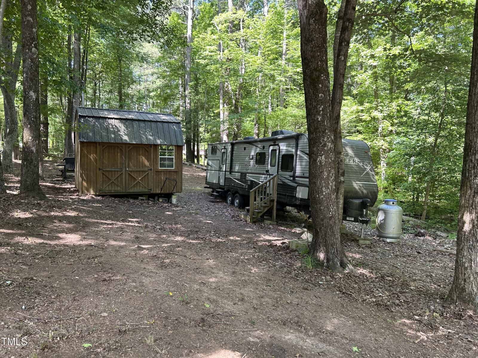 A small wooden cabin and a travel trailer are set among tall trees in a shady, wooded area. The ground is covered with dirt and leaves, and sunlight filters through the green foliage.