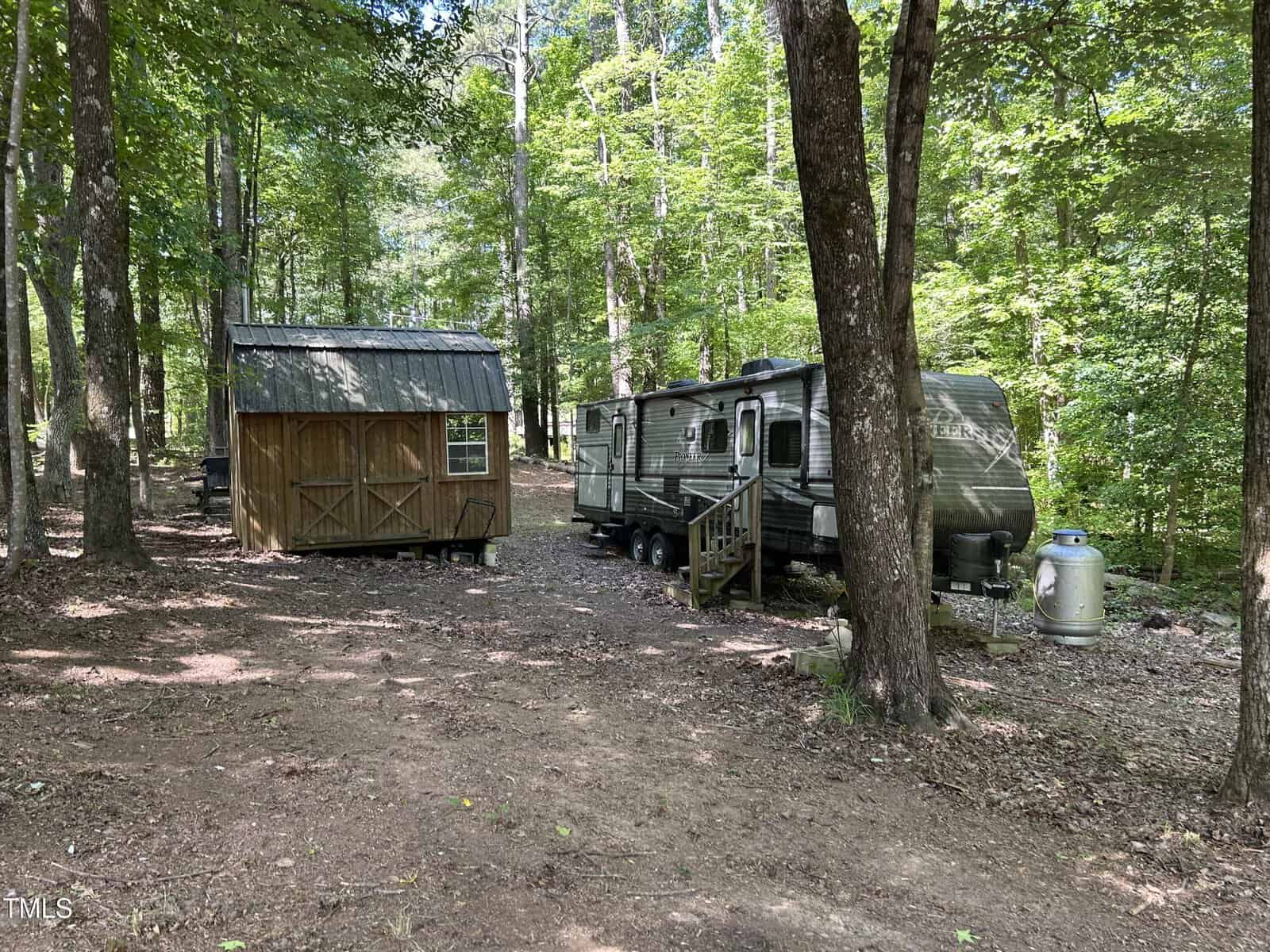 A small wooden shed and a travel trailer sit among tall trees in a sunlit forest clearing, with a propane tank next to the trailer and dappled sunlight on the forest floor.