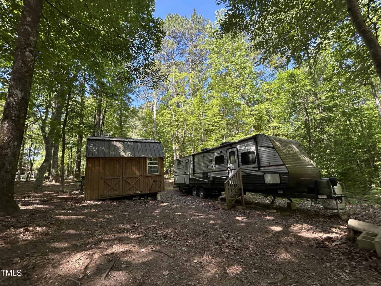 A small wooden cabin and a silver travel trailer sit side by side in a shady, wooded clearing with tall trees and sunlight filtering through the leaves.