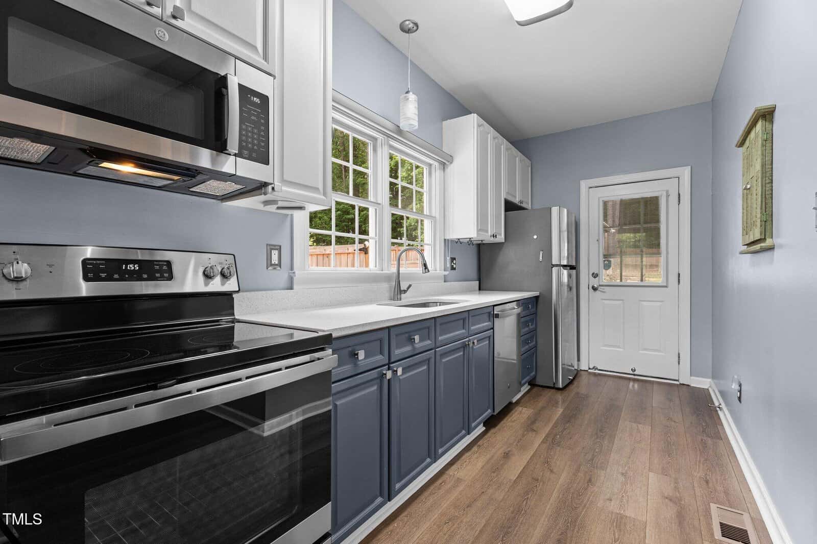 Modern galley kitchen with blue lower cabinets, white upper cabinets, stainless steel appliances, a large window over the sink, light wood floor, and a glass door leading outside.