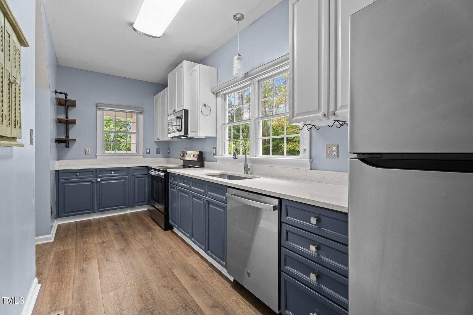 Modern kitchen with blue lower cabinets and white upper cabinets, stainless steel appliances, light wood flooring, large window over the sink, and light blue walls. Natural light brightens the space.